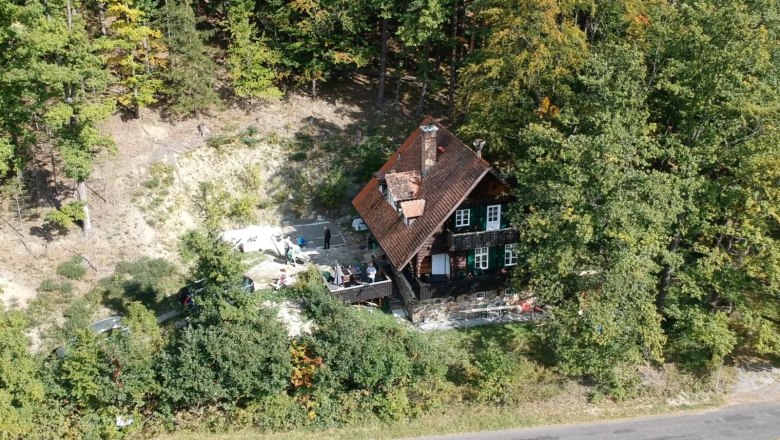 Luftaufnahme eines Hauses im Wald mit rotem Dach und grünen Fensterläden, umgeben von Bäumen.
