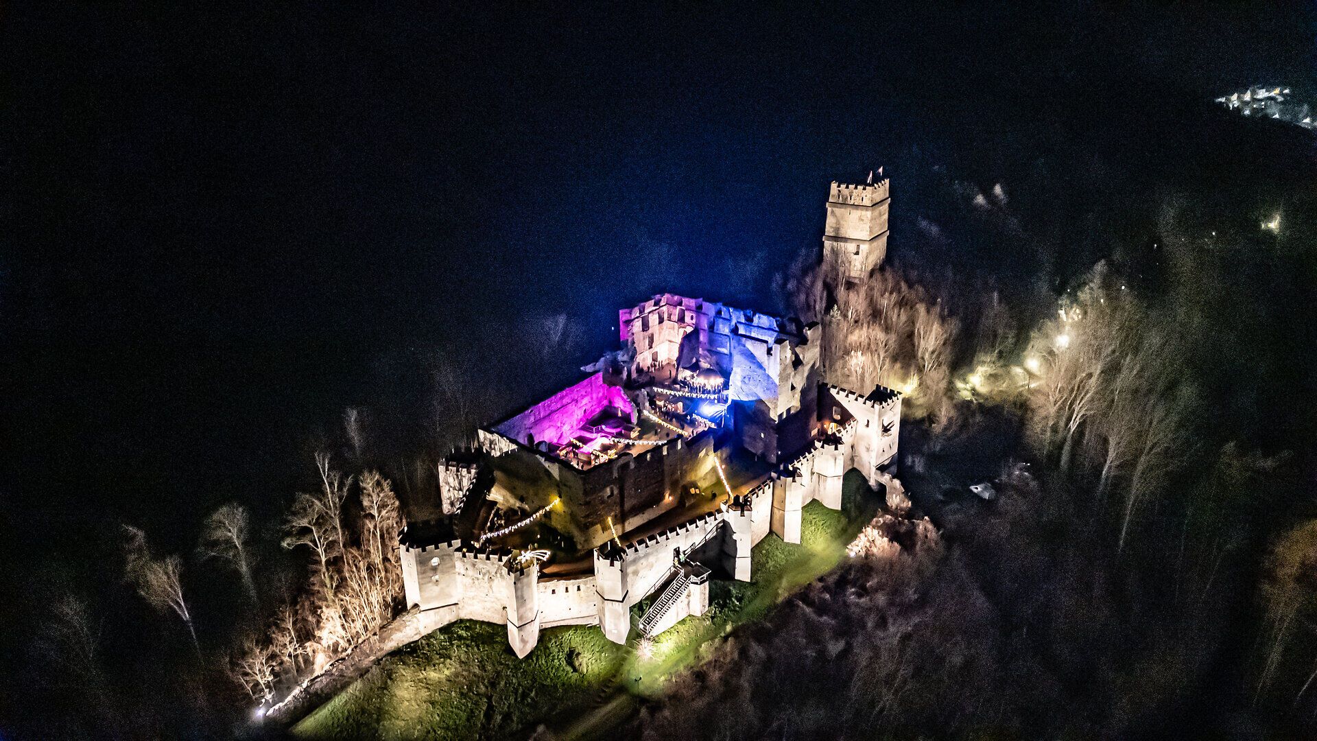 Luftaufnahme einer beleuchteten Burganlage bei Nacht, umgeben von dunklem Wald.