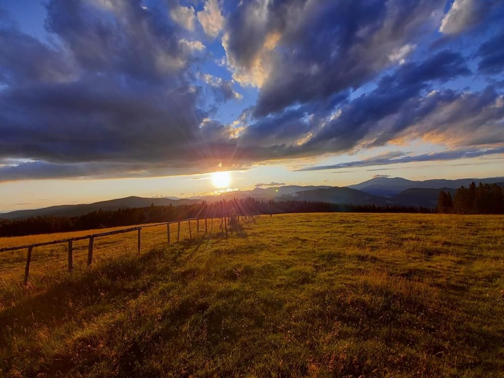 Sonnenuntergang über einer grünen Wiese mit Bergen im Hintergrund und dramatischen Wolken am Himmel.