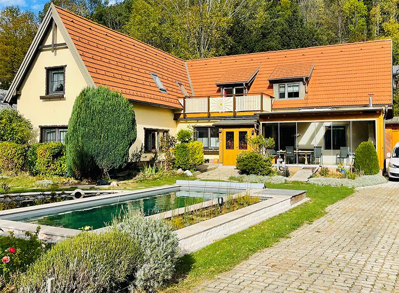 A yellow house with a red roof, garden and small pond in the foreground.