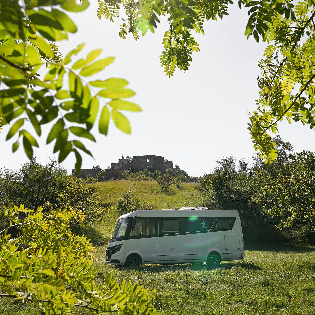 Umgeben von üppigem Grün und unter einem strahlend blauen Himmel, bietet der Campingwagen einen perfekten Rückzugsort für Naturliebhaber. Die sanften Hügel im Hintergrund und die historische Burgruine schaffen eine malerische Kulisse, die zum Entspannen und Erkunden einlädt.