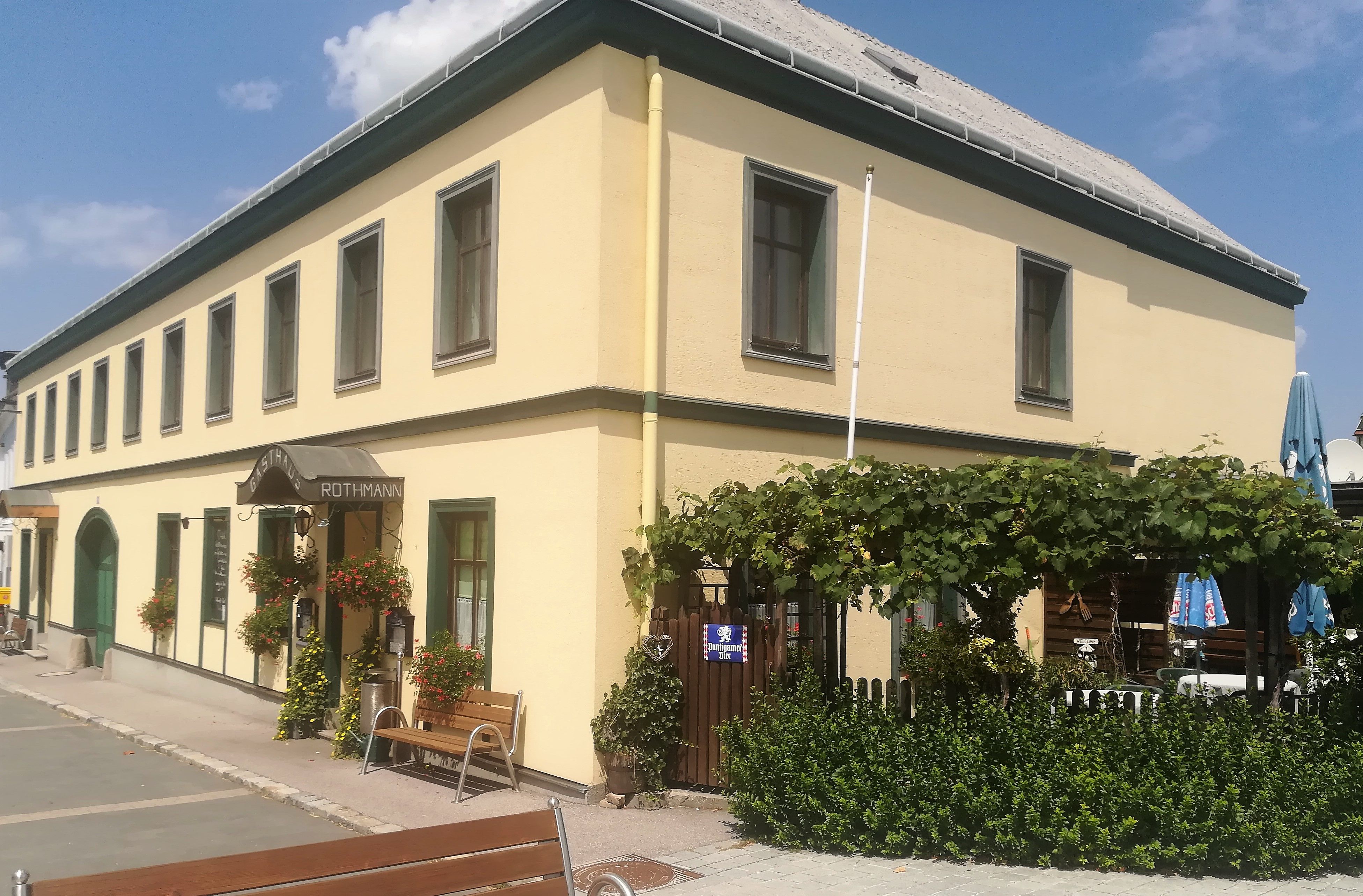 Gelbes Gasthaus mit grünen Fensterrahmen, Gastgarten und Pflanzen vor dem Eingang.