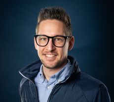 Portraitfoto Wiener Alpen Mitarbeiter, der eine Jacke mit Wiener Alpen Logo trägt und vor einem dunklem Hintergrund steht.