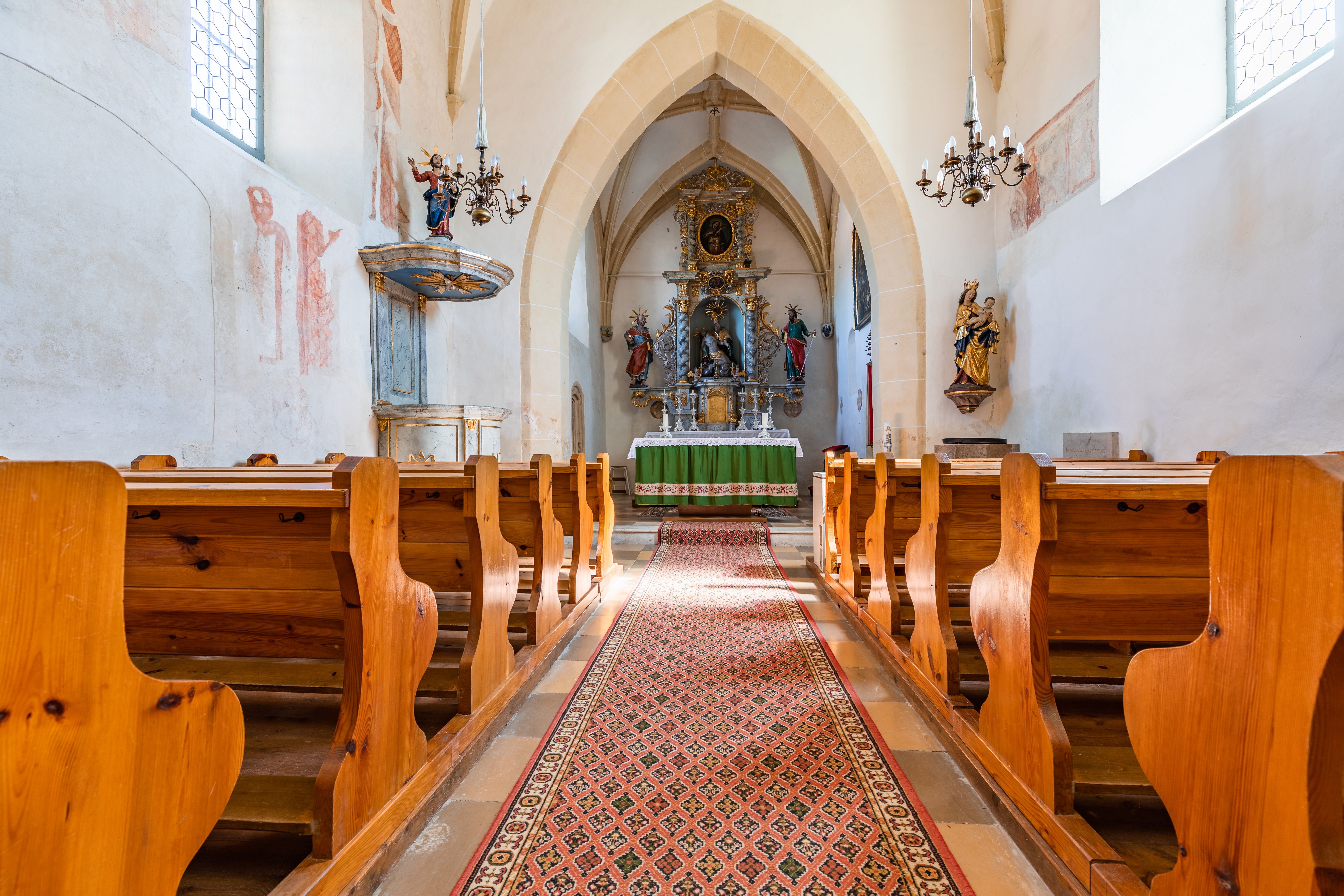 Das Innere der Wehrkirche Bad Schönau, vom Mittelgang aus fotografiert, mit hohen Steinmauern, Holzbänken, Kanzel und Altarraum. An den Wänden verblasste Wandmalerei.