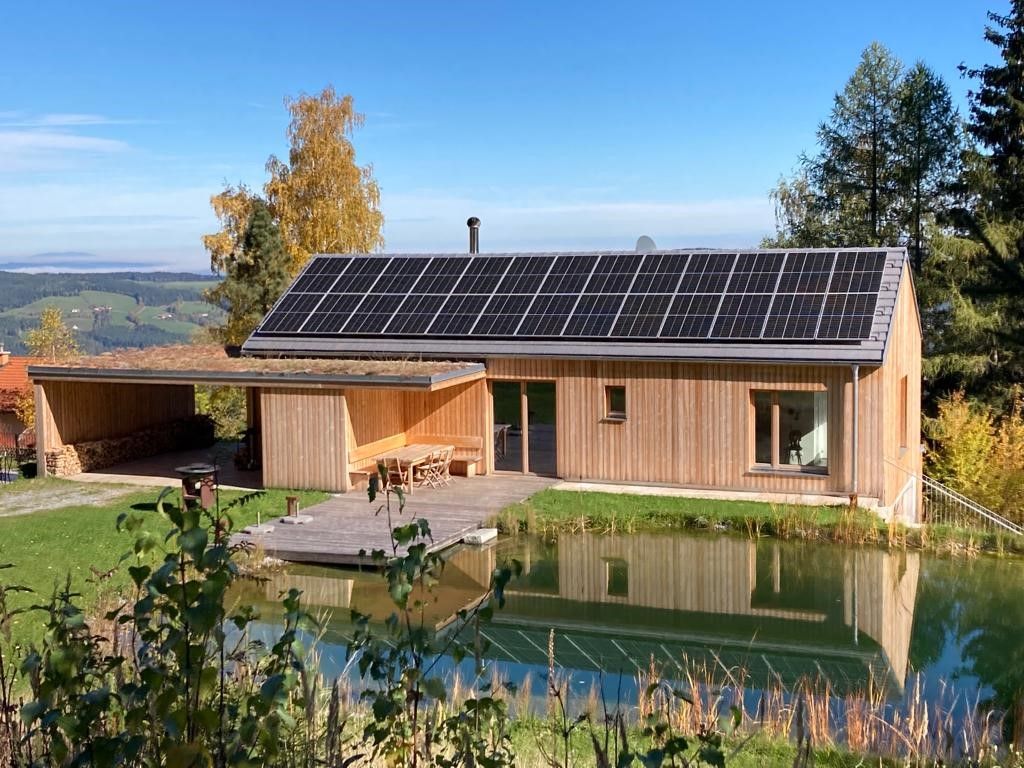 Ein modernes Holzhaus mit Solarpanelen auf dem Dach, umgeben von Natur und einem kleinen Teich im Vordergrund.