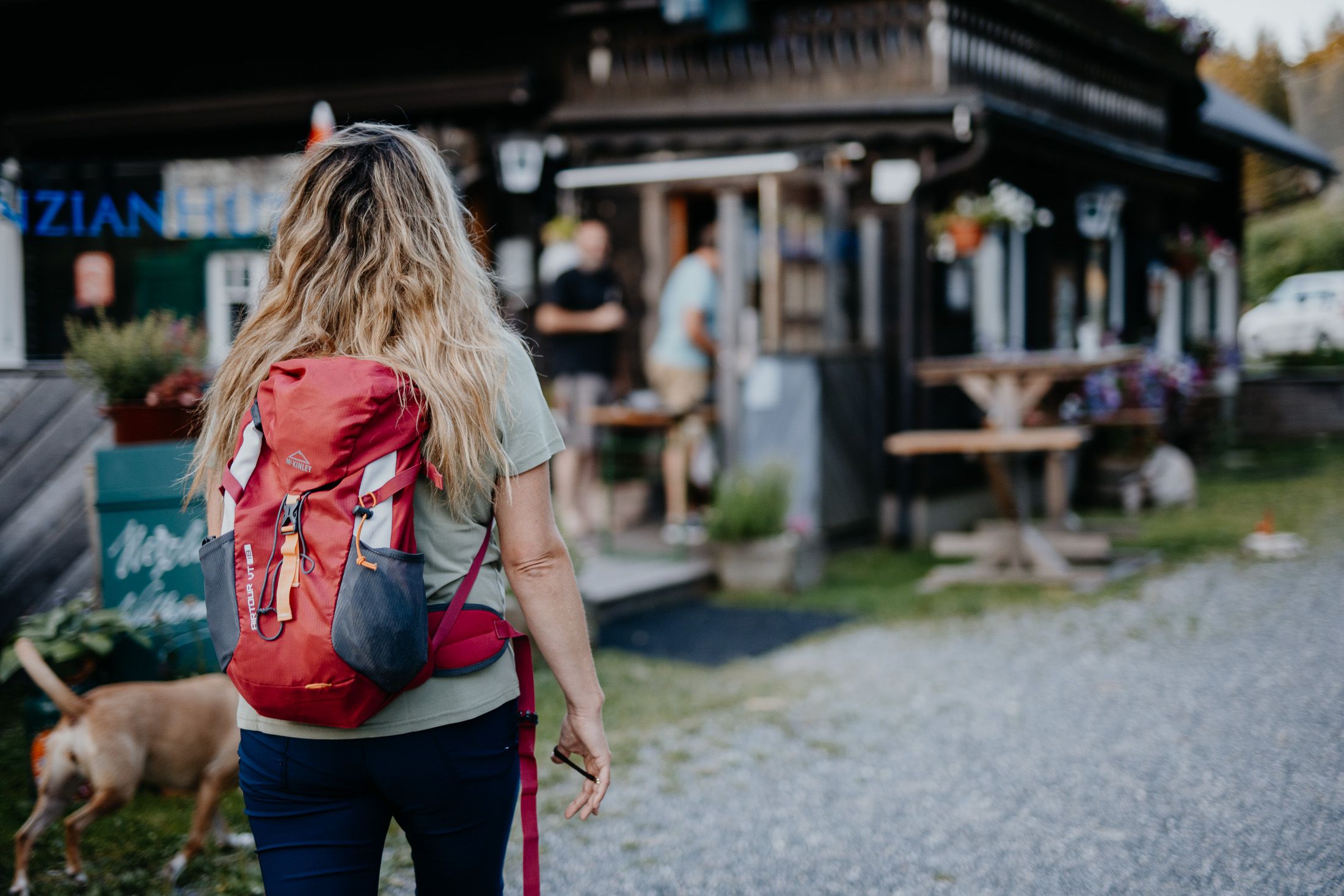 Eine Wanderin mit rotem Rucksack geht auf die Holzhütte zu.