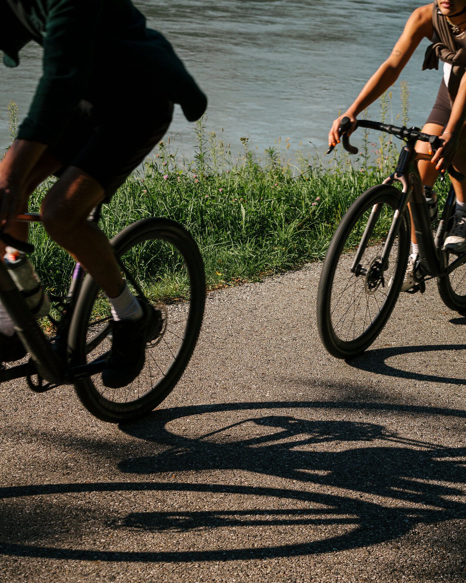 Zwei Radfahrende auf einem asphaltierten Radweg direkt am Fluss; zu sehen sind Räder, Beine und die Schatten auf dem Boden.