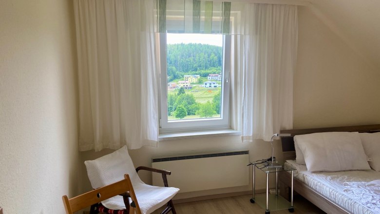 Helles Schlafzimmer mit Fensterblick auf grüne Landschaft, Bett, Stuhl und Tisch.
