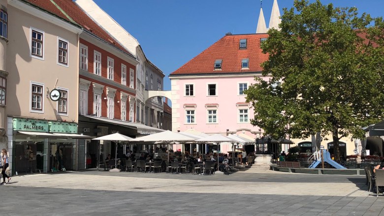 Ein Platz mit Cafés und Geschäften, Sonnenschirmen und einem Baum.