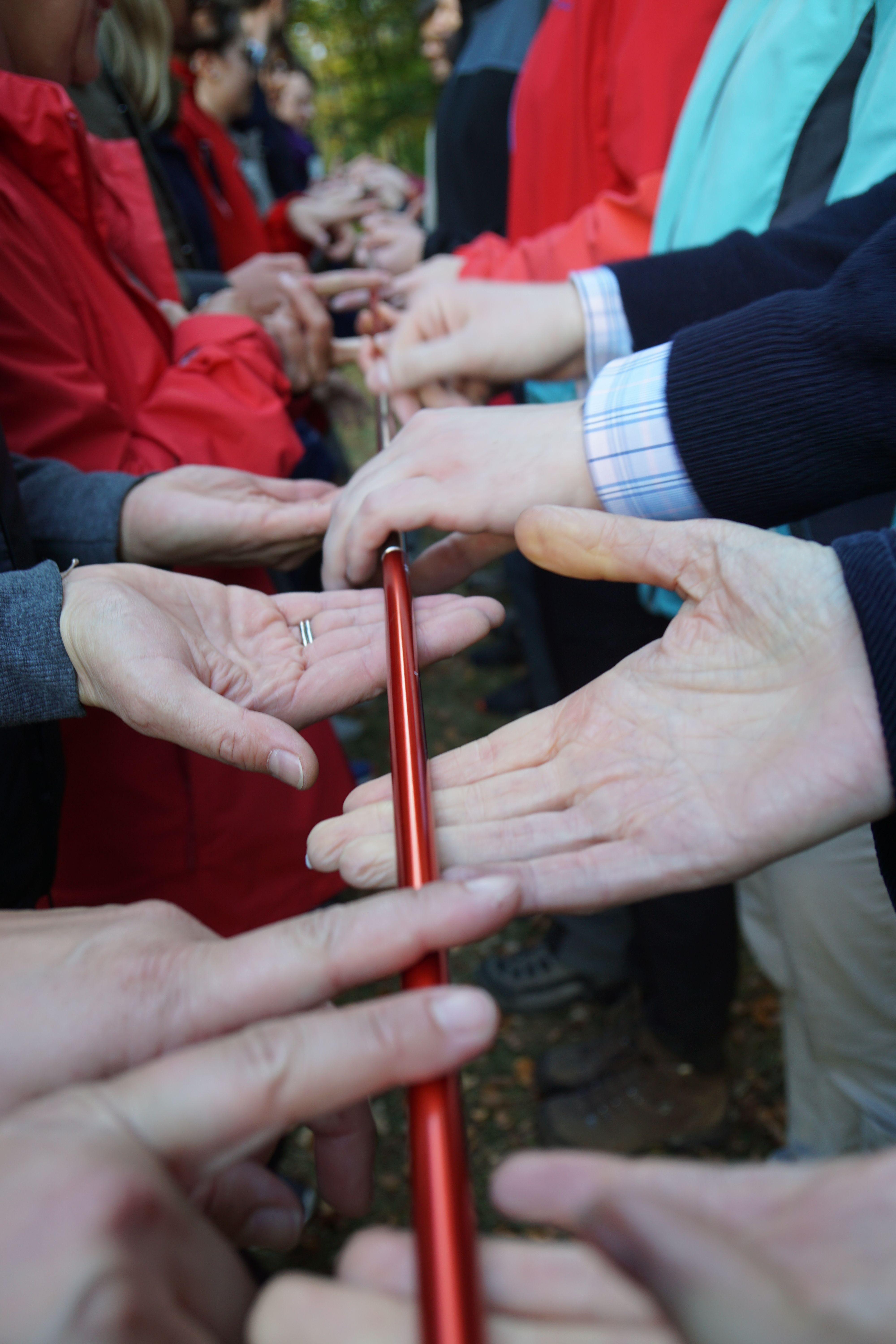 Menschen halten gemeinsam einen roten Stab mit den Fingern.