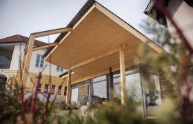 Modern building with wooden roof and glass front, surrounded by plants.