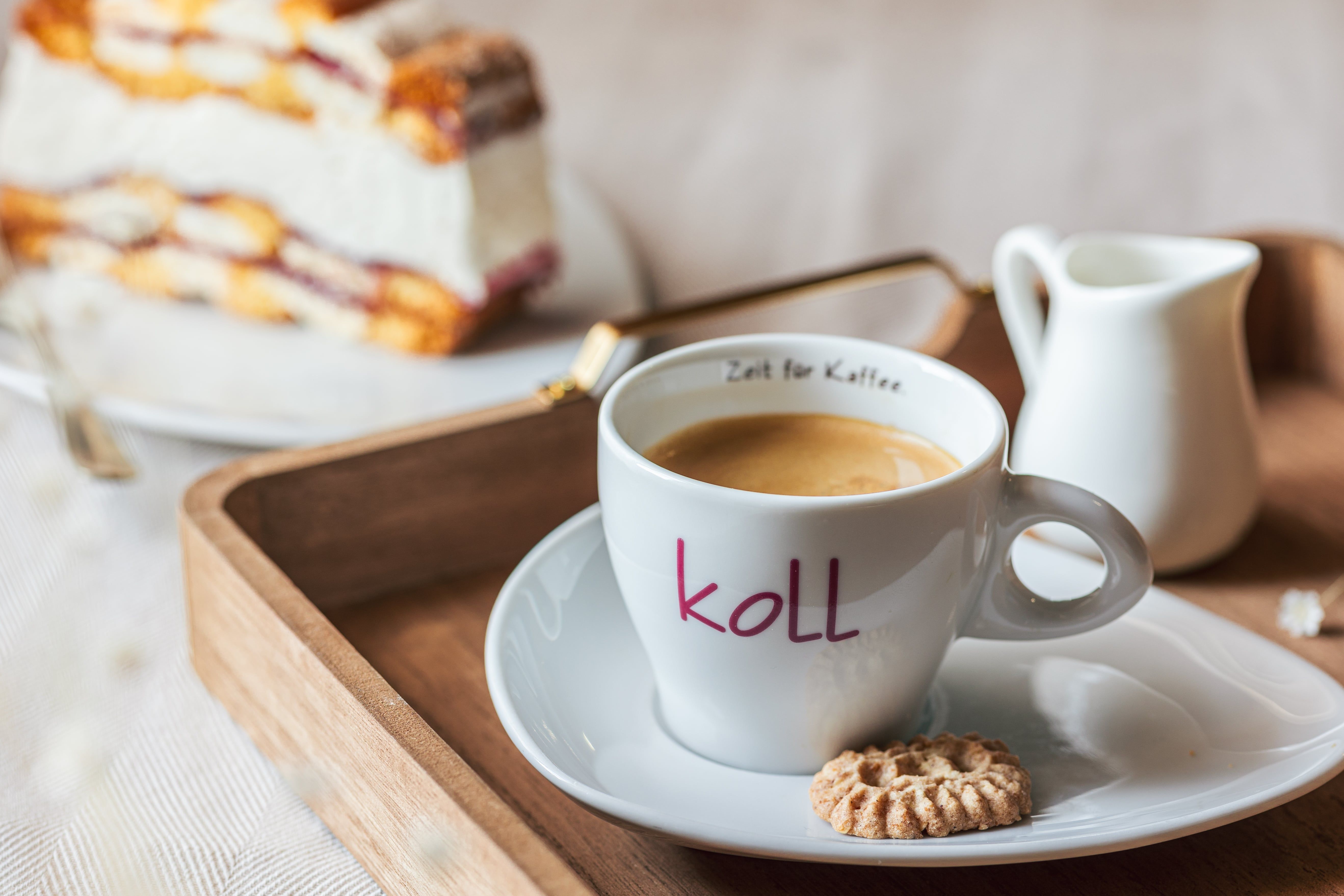 Eine Tasse Kaffee mit einem Keks auf einem Tablett, im Hintergrund ein Stück Torte.
