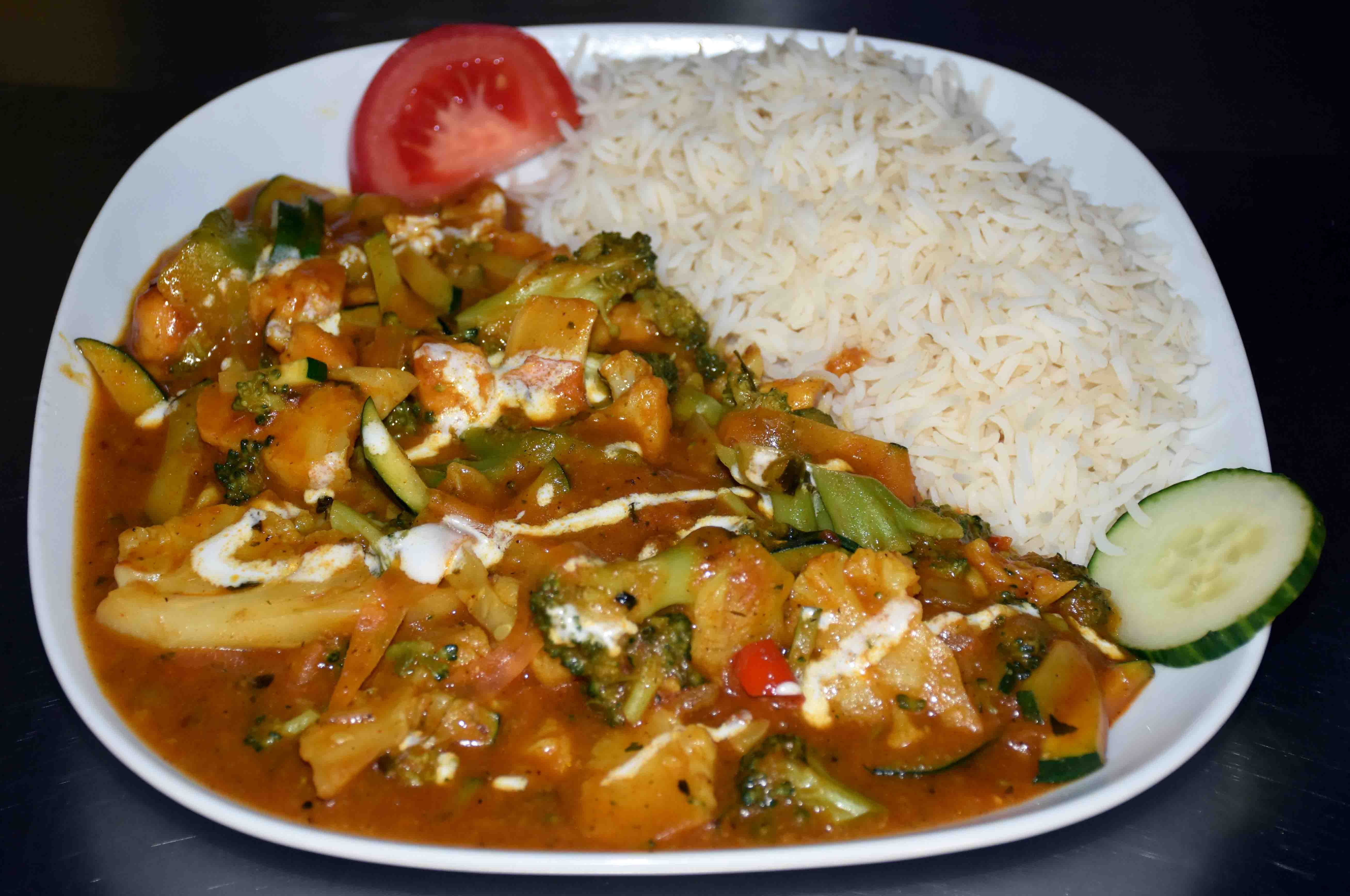 Ein Teller mit einem Gemüsegericht in Soße und Reis, garniert mit einem Stück Tomate und Gurke.