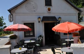 Außenansicht eines Cafés mit Terrasse, Tischen und Sonnenschirmen.