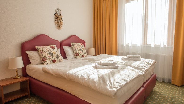 Ein gem&uuml;tliches Doppelzimmer mit zwei Betten, gelben Vorh&auml;ngen und einem Traumf&auml;nger an der Wand.