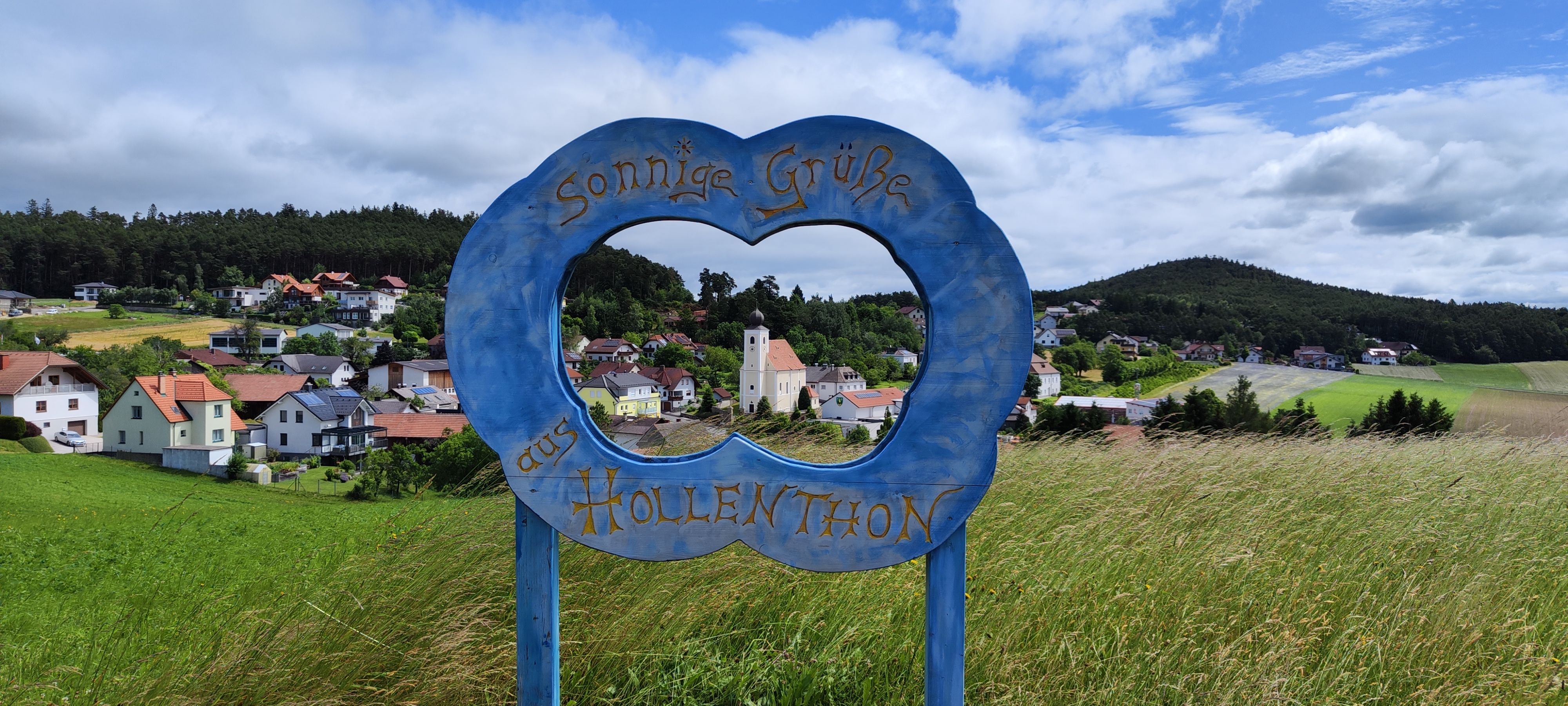 Blick durch einen blauen Rahmen mit der Aufschrift 'Sonnige Grüße aus Hollenthon' auf eine malerische Landschaft mit Dorf und Kirche.