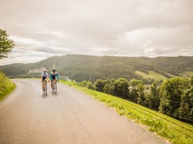 Zwei Personen am Fahrrad auf einer Stra&szlig;e in h&uuml;geliger Landschaft mit einer Burg im Hintergrund.