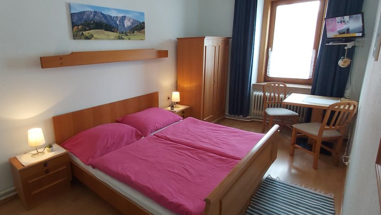 Ein gemütliches Doppelzimmer mit Holzmöbeln, einem Doppelbett mit pinker Bettwäsche, einem Tisch mit zwei Stühlen und einem kleinen Fernseher.