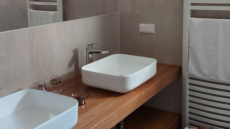 Modernes Badezimmer mit zwei weißen Waschbecken auf Holzplatte, Handtuchhalter und Korb mit Handtüchern.