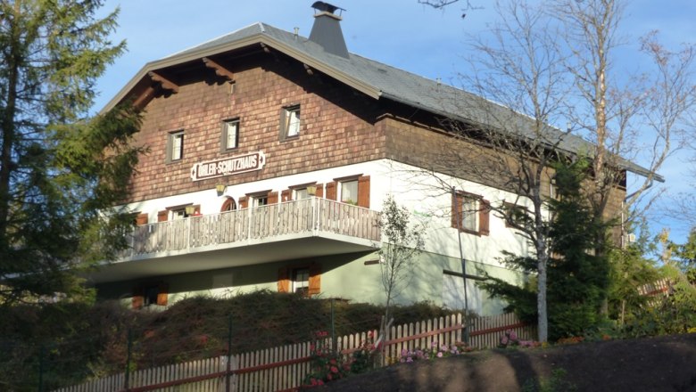 Ein traditionelles Gebäude mit Holzverkleidung und Balkon, umgeben von Bäumen und einem Holzzaun. Schild: "Döhler-Schutzhaus".