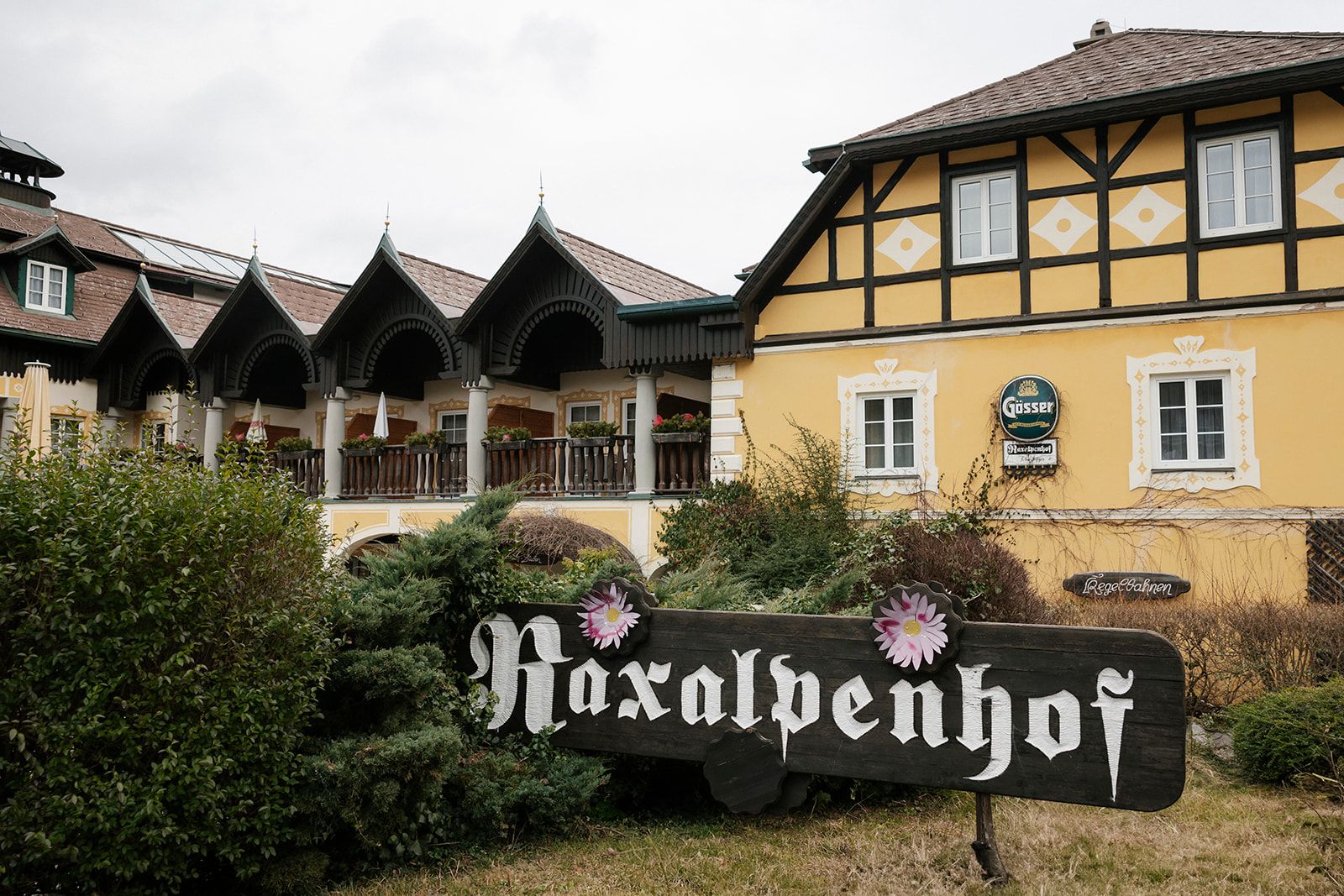Außenansicht des Raxalpenhofes mit gelber Fassade, davor ein großes Holzschild mit der Aufschrift Raxalpenhof