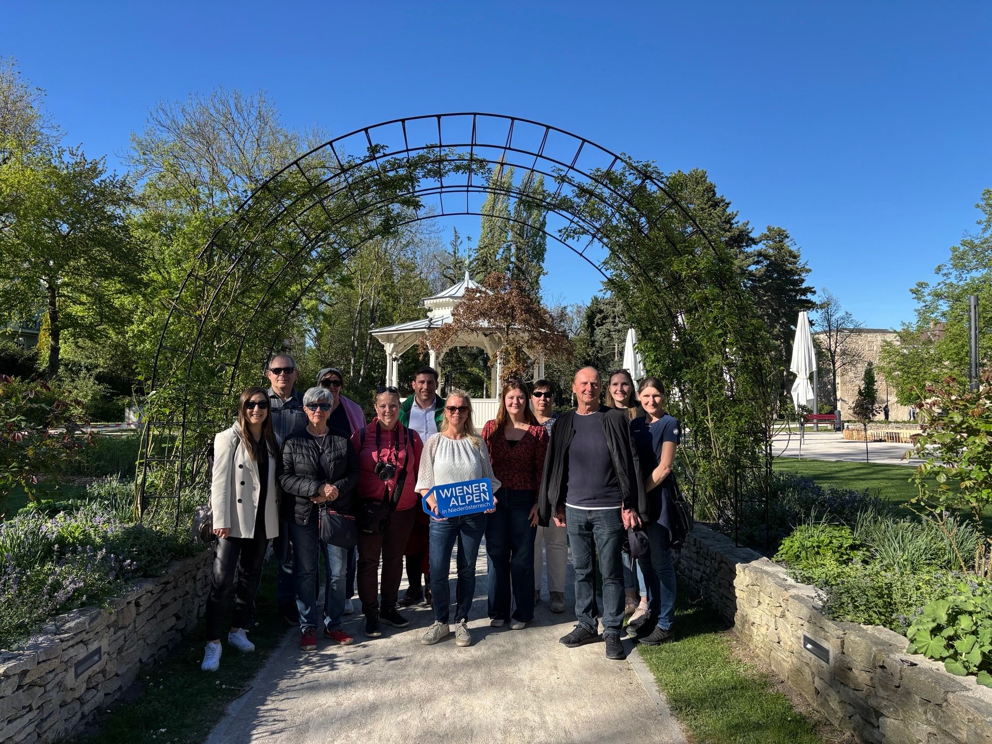 Eine Gruppe von rund 12 Personen steht im Stadtpark Wiener Neustadt vor dem Pavillon und einem Rosenbogen. Eine Frau hält ein Wiener Alpen Logo-Schild in der Hand.