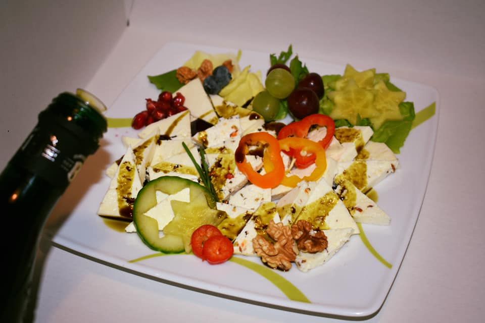 Ein Teller mit Schafkäse, dekoriert mit Paprika, Gurke, Nüssen und Früchten. Eine Flasche mit Öl wird darüber gehalten.