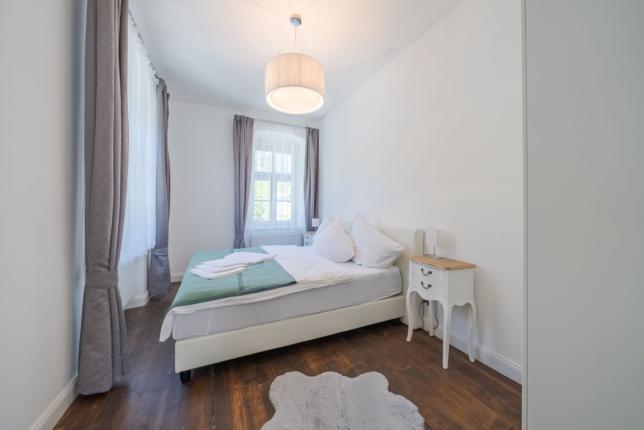Ein helles Schlafzimmer mit einem Doppelbett, weißen Wänden und Holzboden. Ein Nachttisch steht neben dem Bett, und ein großer Lampenschirm hängt von der Decke.