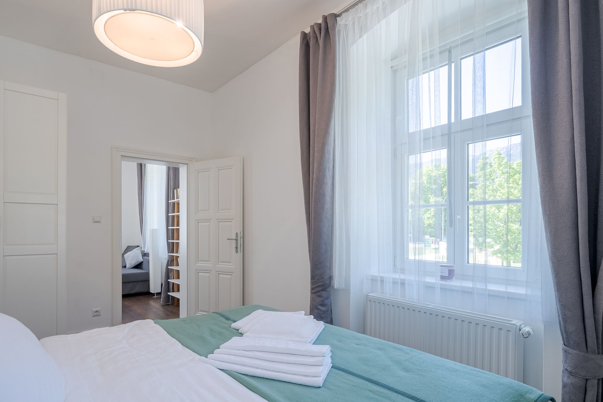 Helles Schlafzimmer mit großem Fenster, weißen Vorhängen und einem Bett mit gefalteten Handtüchern.