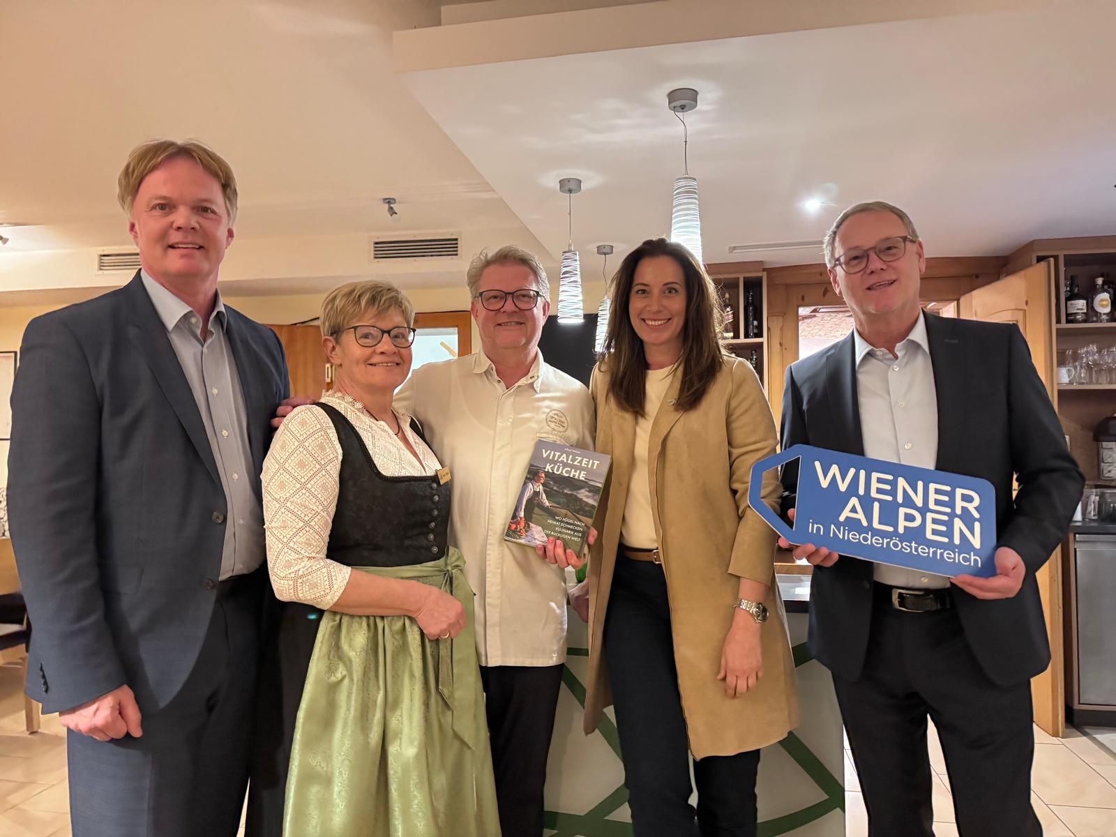 Fünf Personen stehen nebeneinander. Ein Mann hält ein Logoschild in seinen Händen, eine Person hält ein Kochbuch in seinen Händen.