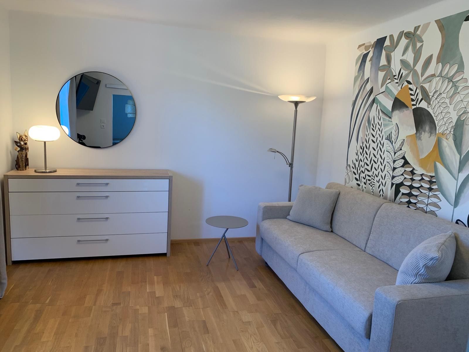 Modernes Wohnzimmer mit grauem Sofa, Kommode, Spiegel und dekorativer Wandkunst.