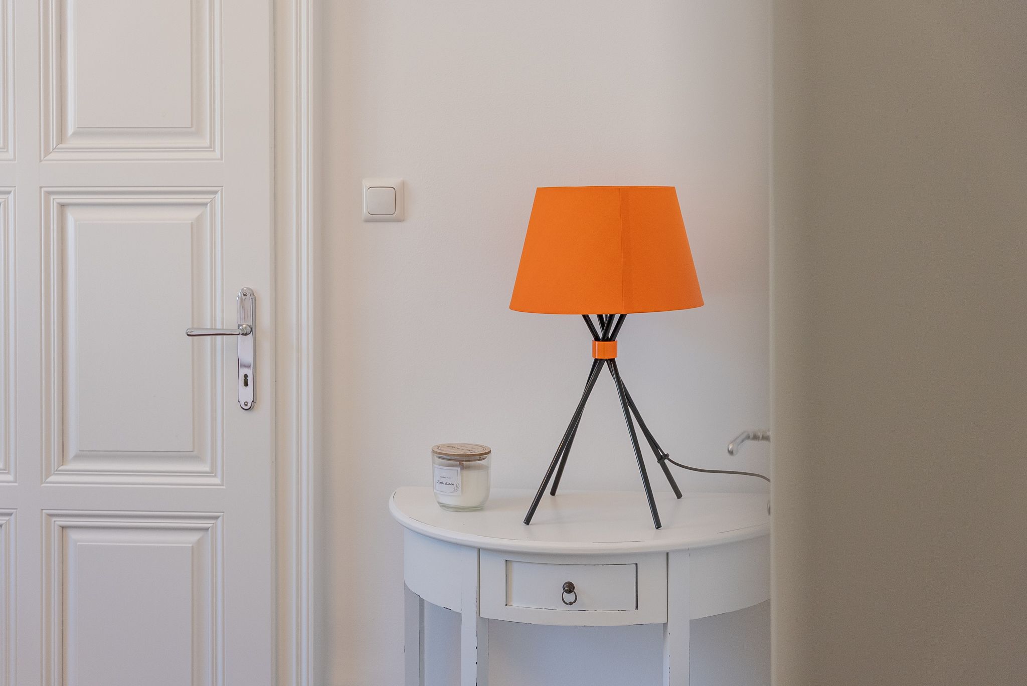 Ein weißer Tisch mit einer orangefarbenen Lampe und einer Kerze darauf, neben einer weißen Tür.