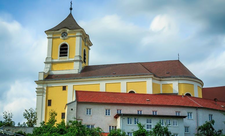 Gelbe Kirche mit Turm und roten Dächern vor bewölktem Himmel.