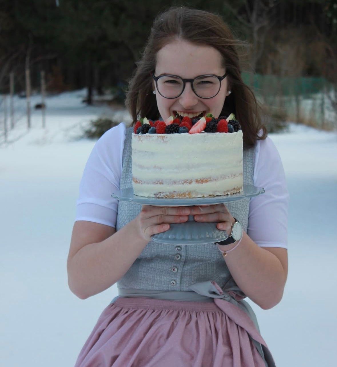Eine lächelnde Frau mit Brille hält im Schnee eine mit Beeren verzierte Torte auf einer Glasplatte.