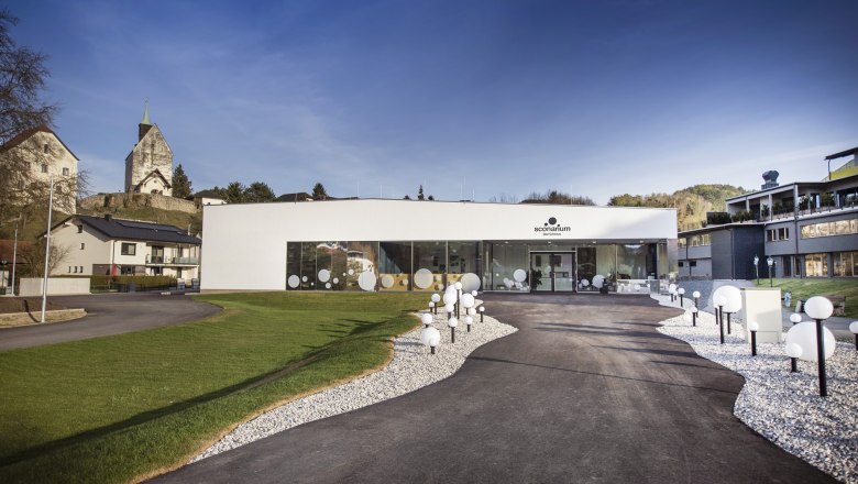 Modernes Geb&auml;ude des Sconariums mit Glasfront und dekorativen Kugelleuchten, umgeben von Gr&uuml;nfl&auml;chen und der Wehrkirche im Hintergrund.