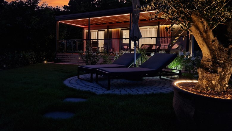 Exterior view of a chalet at sunset with sun loungers and illuminated tree in the foreground.