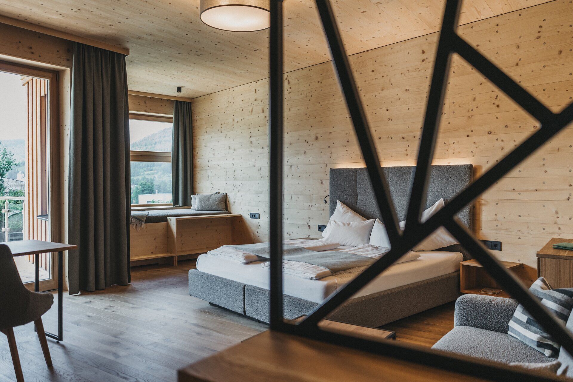Ein gemütliches Zimmer, dessen Wände und Decke komplett aus hellem Naturholz gefertigt sind. Ein graues Polsterbett steht vor der Holzwand; im Hintergrund ist eine in die Wand integrierte Sitznische am Fenster erkennbar. Das Bild wird durch ein im Vordergrund unscharf eingeblendetes, dunkles Dekoelement eingerahmt.
