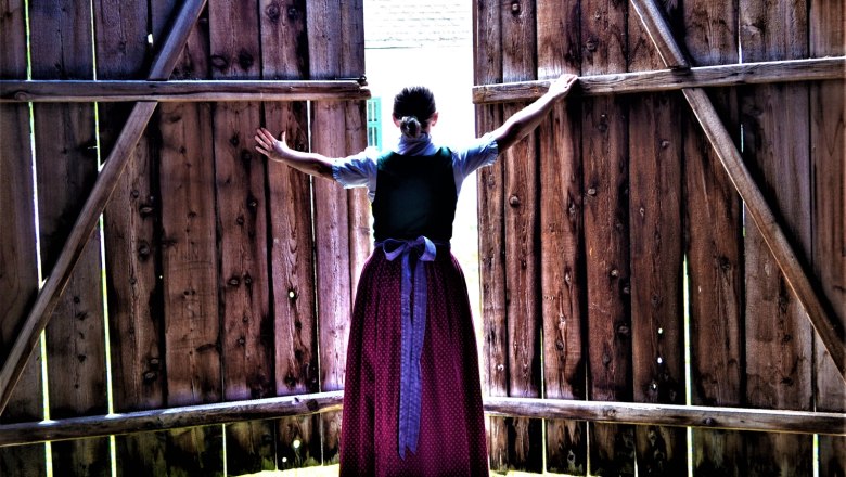 Eine Frau in traditioneller Kleidung öffnet große Holztüren nach außen.