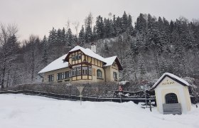 Winterliche Villa Waldheimat mit verschneitem Dach und Wald im Hintergrund.