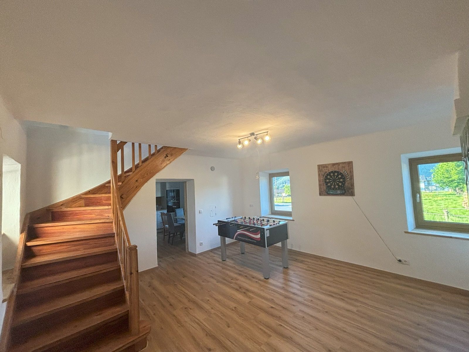 Ein Gemeinschaftsraum mit Tischfußballtisch, Dartscheibe an der Wand, Holztreppe und zwei Fenstern.