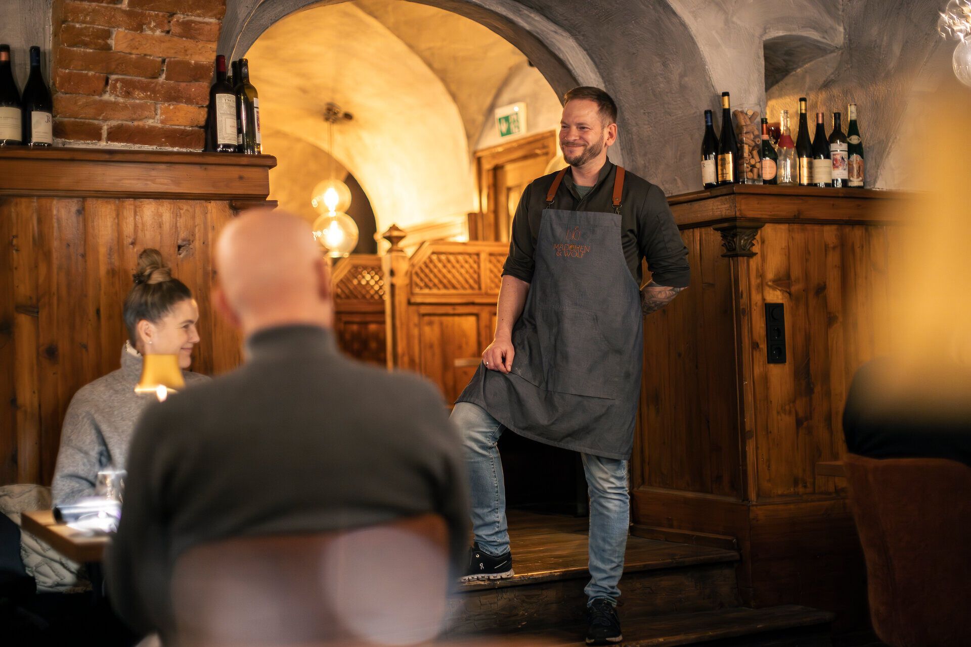 Ein Mann mit Schürze steht mit einem Fuß auf einer Stufe lächelnd vor Gästen in einer Gaststube.