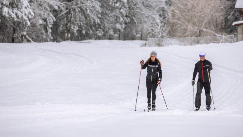 Zwei Personen beim Langlaufen auf einer verschneiten Loipe, umgeben von schneebedeckten Bäumen.