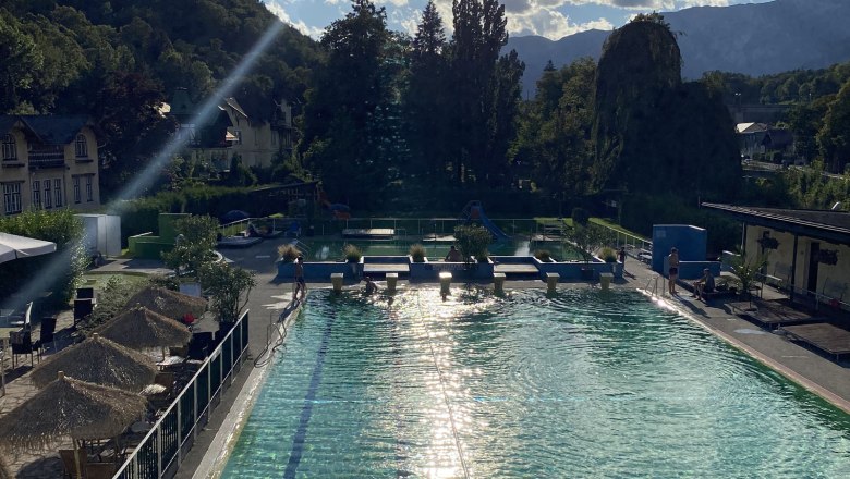 Ein Freibad mit klarem Wasser, umgeben von Bäumen und Bergen im Hintergrund.