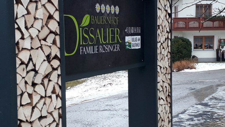 Schild eines Bauernhofs mit Holzstapeln und einem Haus im Hintergrund.