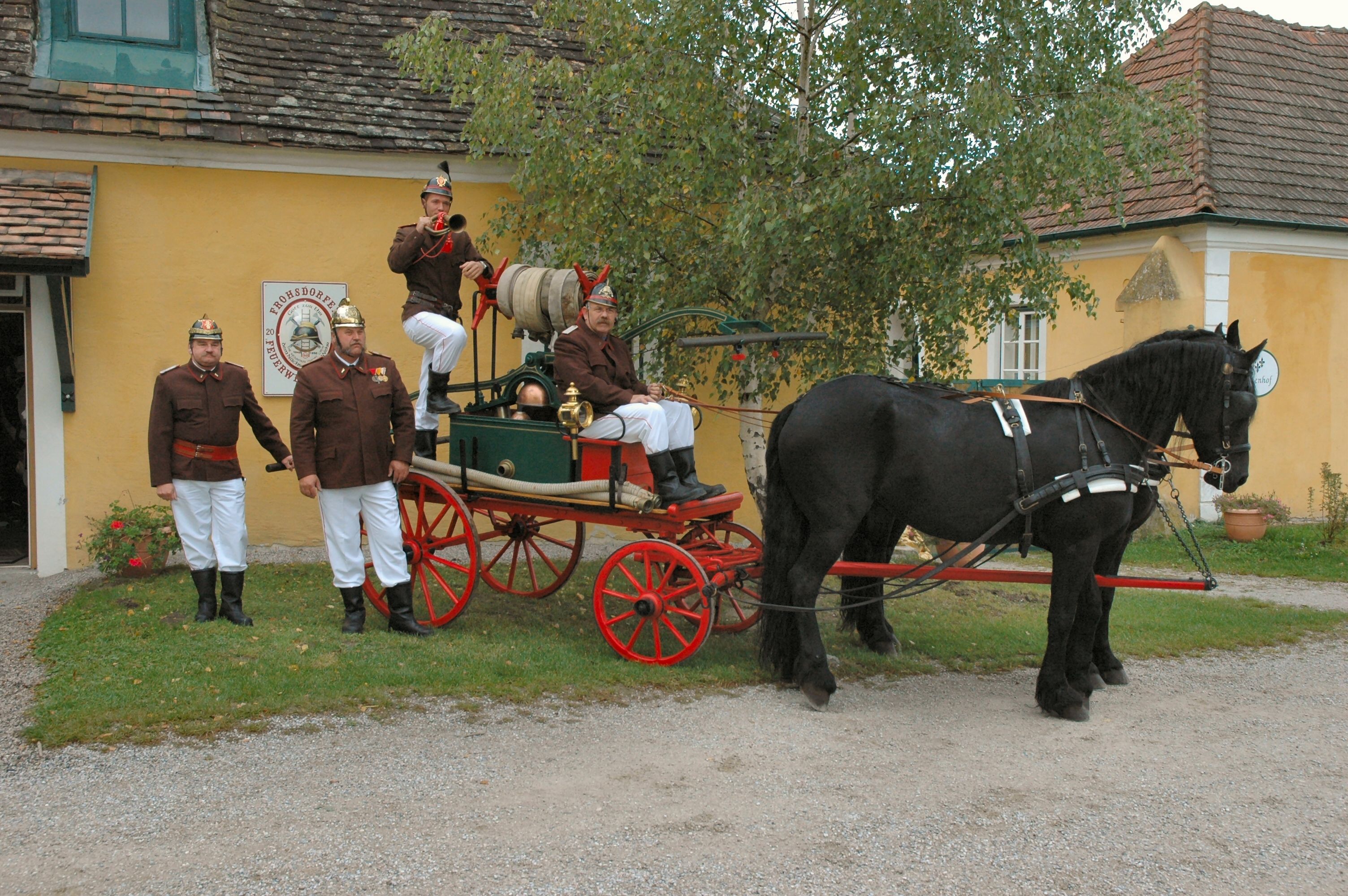 Historische Feuerwehrkutsche mit Pferd und Feuerwehrleuten in Uniform vor einem gelben Gebäude.