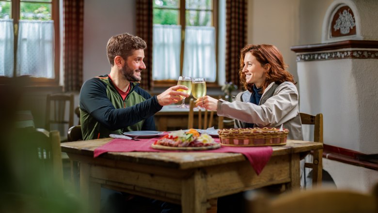 A couple sits in a parlor on a rustic wooden table in front of a Brettljause and toasts with apple cider