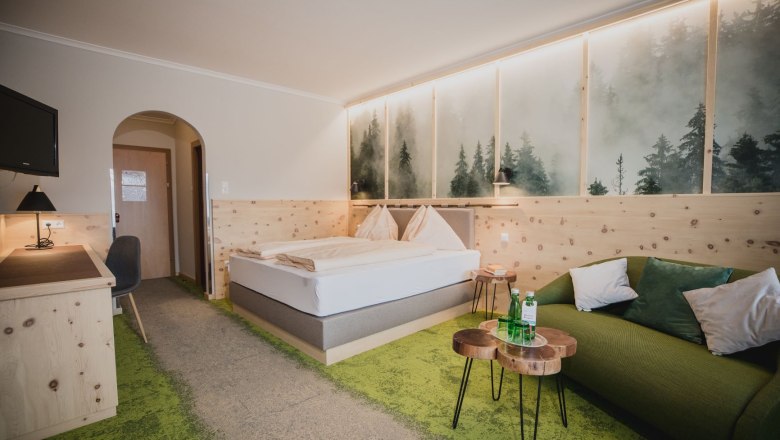 Ein gemütliches Hotelzimmer mit Zirbenholzmöbeln, einem Doppelbett, grünem Teppich und einem Sofa. An der Wand ein Bild von nebligem Wald.