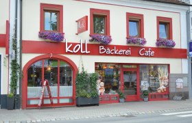 Außenansicht der Koll Bäckerei Café in Kirchschlag mit roten Fensterrahmen und Blumenkästen.