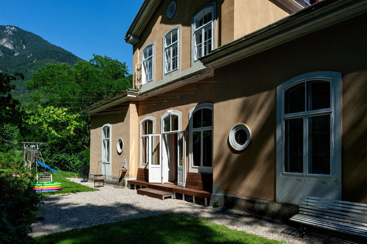Ein zweistöckiges Haus mit großen Fenstern und einem Garten mit Spielplatz vor einer Bergkulisse.