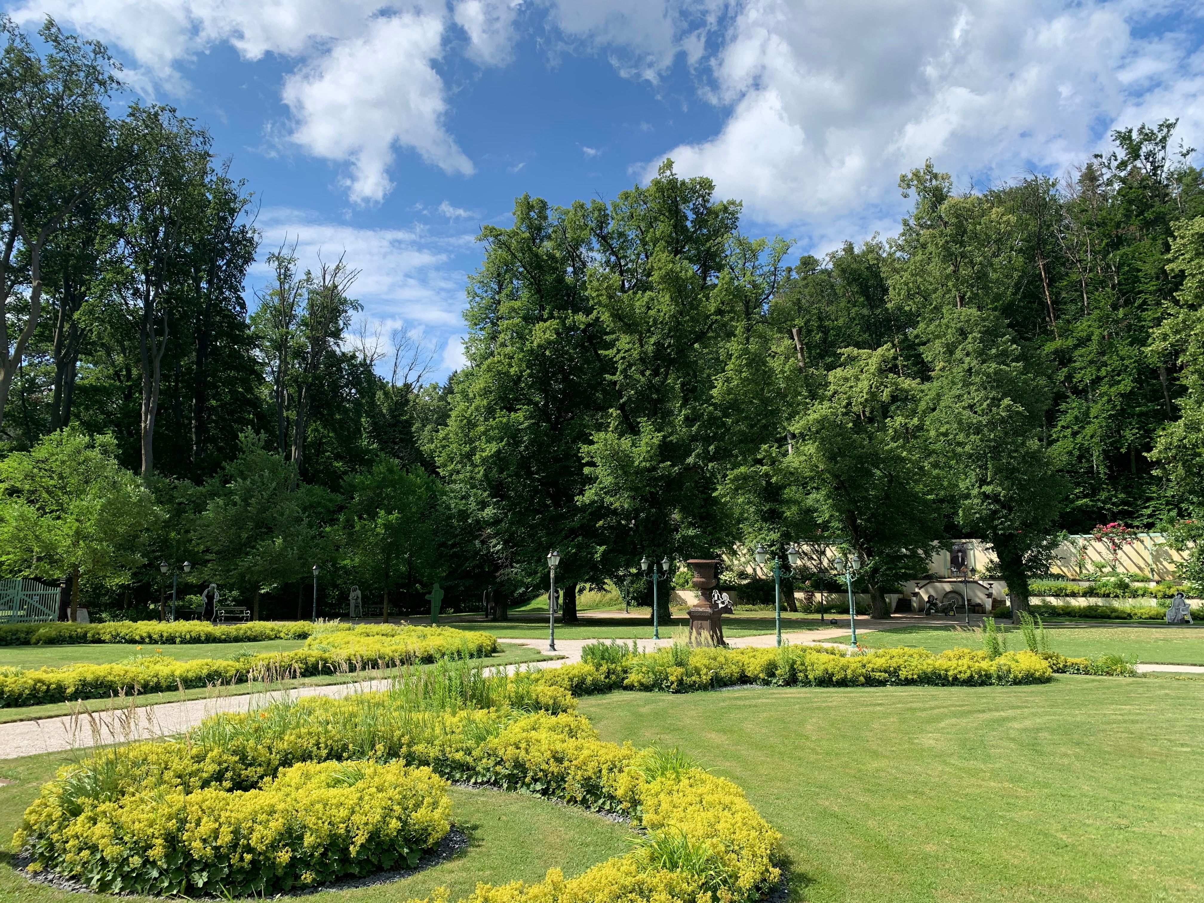 Ein gepflegter Garten mit grünen Bäumen und gelben Blumenbeeten unter einem blauen Himmel mit Wolken.